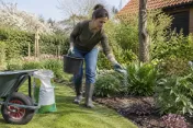 Vrouw strooit kunstmest uit met emmer en handschoenen in bloementuin tijdens daglicht